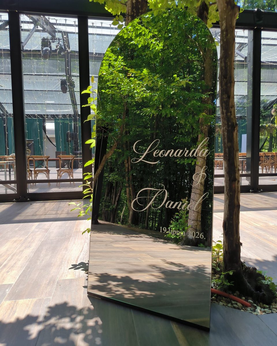 Lučno ogledalo dobrodošlice s imenima Leonarda & Daniel ispred staklenog paviljona, s drvećem koje se zrcali u staklu.