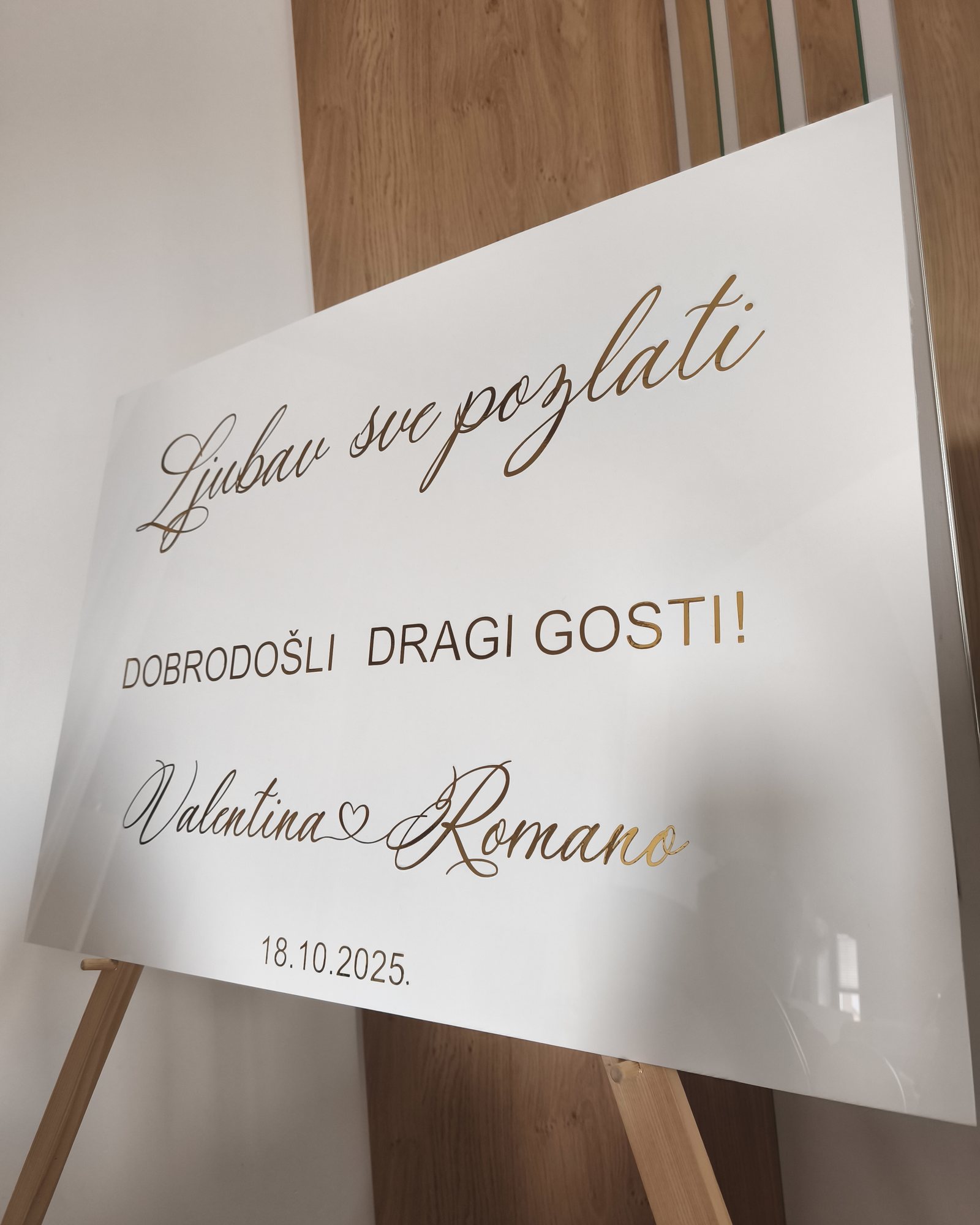 Bijela ploča na drvenom stalku sa zlatnim natpisom „Ljubav sve pozlati“, „Dobrodošli dragi gosti“ i imenima Valentina & Romano, 18. 10. 2025.